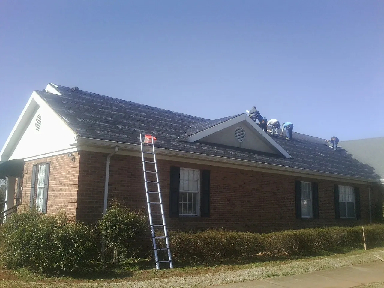 Crew tearing shingles off a brick ranch roof with a ladder set — Metal Roof Repair work by our crew in Belding