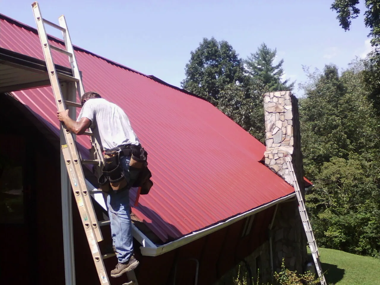 Roofer on a ladder beside a red metal roof and stone chimney — Soffit & Fascia Repair by Roofing Brothers in Belding