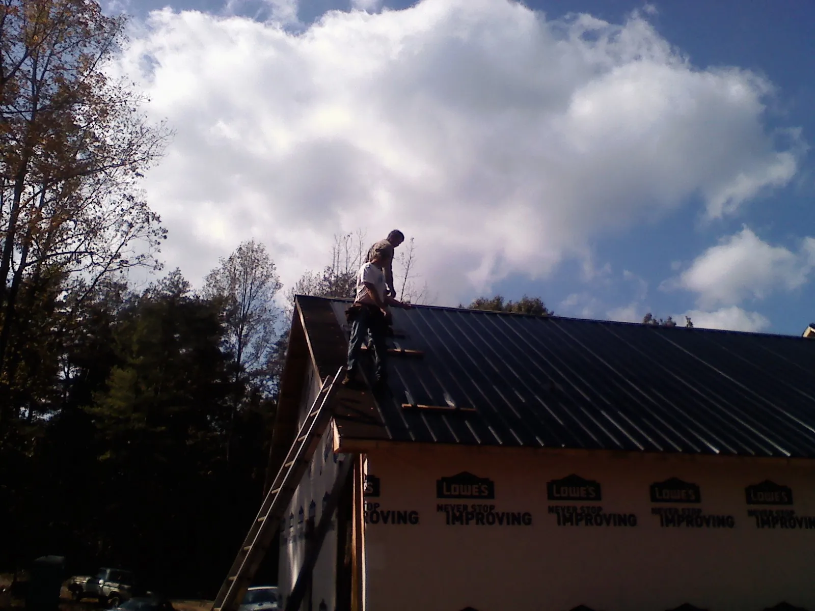 Roofer fastening dark metal panels high against a cloudy sky, from a Roof Inspection job in Belding